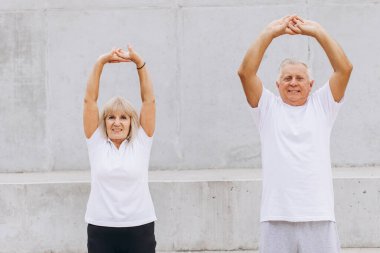 Sağlıklı bir yaşam tarzı için dışarıda esneme egzersizleri yapan kıdemli bir çift. Beyaz gömlekli bir erkek ve bir kadın spor ve sağlık aktivitelerinden zevk alır. Huzurlu bir ortamda yaşlılar arasında aktif yaşamı teşvik etmek.