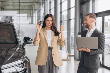 Yeni arabanın anahtarlarını tutan, yumruğunu gösterip modern araba galerisinde evet diyen beyaz bir kadın. Mutlu alıcı kız rüyasını gerçekleştirdikten sonra sevinir..