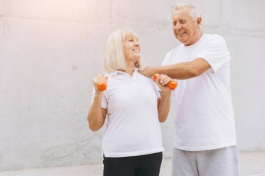 Üst düzey bir çift açık havada parlak turuncu halterlerle egzersiz yapıyor. Görüntü pozitif ve sağlıklı bir yaşam tarzını, uygunluğu, yaşlılığı ve refahı tasvir ediyor. Onların neşeli ifadeleri birlikte aktif kalmanın sevincini gösteriyor..