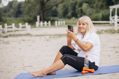 Sahilde spor yapan yaşlı bir kadın, online antrenman için akıllı telefon kullanıyor. Doğada aktif yaşam tarzı, teknoloji ve refah kavramı.