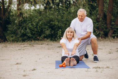 Üst düzey çiftler yoga egzersizlerine katılıyorlar. Doğal ortamlarında spor, sağlık ve beraberlik gösterisi yapıyorlar. Bir erkek, bir kadının egzersiz yapmasına yardım eder..