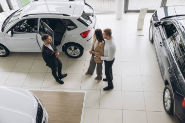Araba galerisinde yeni araba seçen çiftin üst görüntüsü, genç adam ve kadının erkek satış müdürüyle konuşurken kucaklaşması, güzel beyaz bir otomobilin yanında durması,