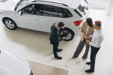 Araba galerisinde yeni araba seçen çiftin üst görüntüsü, genç adam ve kadının erkek satış müdürüyle konuşurken kucaklaşması, güzel beyaz bir otomobilin yanında durması,