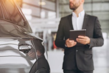 Araba ve araba satıcısına odaklan. Gri takım elbiseli, araba kuaföründe duran ve elinde tablet tutan adama odaklan. Doğru yerde araba almak her zaman zevktir..