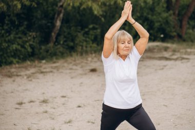 Parkta yoga yapmaktan hoşlanan yaşlı bir kadın gülümseyerek aktif ve sağlıklı bir yaşam tarzı sergiliyor..