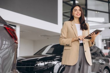 Gülümseyen araba satıcısı, bej takım elbiseli iş kadını araba salonunda duruyor ve doğru arabayı seçmek için tablet kullanıyor. Arka planda birçok modern araba var..