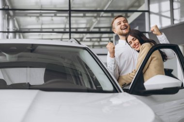 Gayri resmi giyinen mutlu genç çift, galeride ilk yeni aile arabasını alırken sarılıyor. Adam galeride arabanın anahtarlarını gösteriyor..