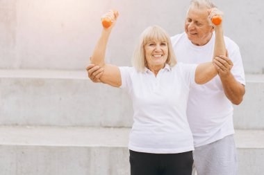 Enerjik yaşlı çift spor yapıyor, dambılları kaldırıyor ve dışarıda gülümsüyor. Aktif bir yaşam tarzını, sağlığını ve altın yıllarında formda ve sağlıklı kalmanın önemini destekliyorlar..