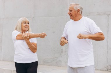Üst düzey bir çift dışarıda egzersiz yapıyor, vücut geliştirme rutinlerinin bir parçası olarak kollarını esnetiyorlar. Günlük spor kıyafetleri giyiyorlar ve sağlıklı bir aktivite ve arkadaşlık anlarını paylaşıyorlar..