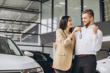 Günlük giysiler içinde mutlu genç bir çift araba galerisinde yeni arabanın anahtarlarını gösteriyor ve sarılıyorlar.