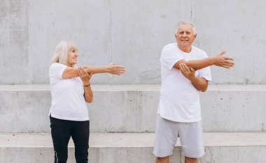 Yaşlı bir çiftin dışarıda egzersiz yaptığı, kollarını aynı anda gerdiği gösteriliyor. İkisi de atletik kıyafetler giyip, spor, sağlık ve altın yıllarında aktif bir yaşam tarzını destekliyorlar..