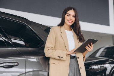 Gülümseyen araba satıcısı, bej takım elbiseli iş kadını araba salonunda duruyor ve doğru arabayı seçmek için tablet kullanıyor. Arka planda birçok modern araba var..