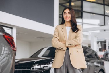 Araba Bayilik ziyaret etti. Güzel genç kadın kameraya bakarak ve çapraz kolları olan dururken yeni arabasını gülümseyen