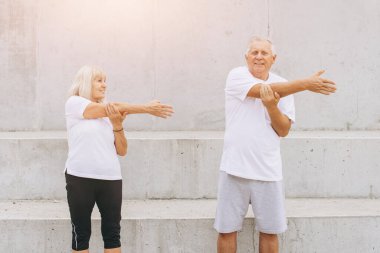 Yaşlı bir çiftin dışarıda egzersiz yaptığı, kollarını aynı anda gerdiği gösteriliyor. İkisi de atletik kıyafetler giyip, spor, sağlık ve altın yıllarında aktif bir yaşam tarzını destekliyorlar..