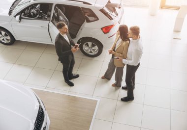 Araba galerisinde yeni araba seçen çiftin üst görüntüsü, genç adam ve kadının erkek satış müdürüyle konuşurken kucaklaşması, güzel beyaz bir otomobilin yanında durması,