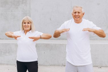 Yaşlı bir çift açık havada spor kıyafetleriyle spor yaparken görülüyor, fitness ve sağlıklı bir yaşam tarzını destekliyorlar. Her ikisi de beyaz gömlek giyiyor ve aktif olarak spor yapıyorlar, pozitiflik sergiliyorlar..