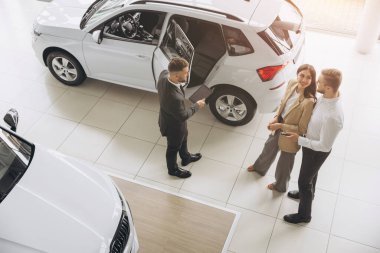 Araba galerisinde yeni araba seçen çiftin üst görüntüsü, genç adam ve kadının erkek satış müdürüyle konuşurken kucaklaşması, güzel beyaz bir otomobilin yanında durması,