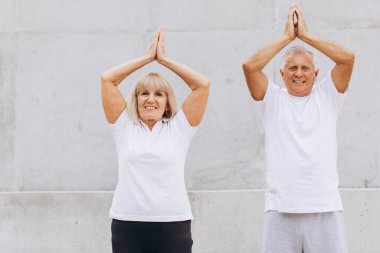 Üst düzey bir çift dışarıda beyaz giysiler içinde yoga yapıyor, sağlık ve sağlığa odaklanıyor. Yaşlı çift, elleri havada yoga pozu vererek sağlıklı ve aktif bir yaşam tarzını destekliyor..