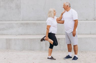 Yaşlı bir çift açık hava fitness rutinine katılıyor. Erkek ona destek olurken kadın bacağını esnetiyor. İkisi de gülümsüyor ve günlük kıyafetlerini giyiyorlar..