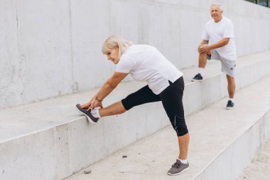 Dışarıda esneme egzersizi yapan yaşlı bir çift. Kadın ayağını beton bir çıkıntıya uzatırken erkek arka planda geriniyor. İkisi de spor kıyafetleri giymiş..