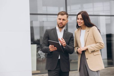 Gri takım elbiseli sakallı iş adamı araba satıcısı araba galerisinin önünde bej ceketli güzel bir bayana tablet üzerinde açıklama yapıyor.