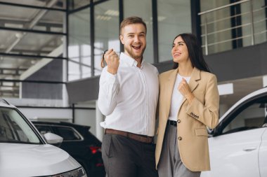 Gayri resmi giyinen mutlu genç çift, galeride ilk yeni aile arabasını alırken sarılıyor. Adam galeride arabanın anahtarlarını gösteriyor.