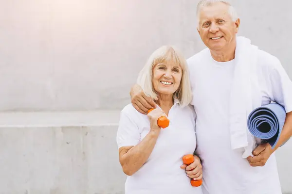 stock image Cheerful senior couple enjoying outdoor exercise with dumbbells and yoga mat. They are wearing white attire and showing healthy lifestyle, fitness, and wellness. Perfect for promoting senior health and active living.