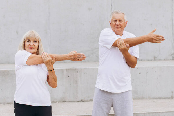 Senior couple engaging in outdoor exercise, stretching their arms to maintain a healthy lifestyle. They are both dressed in comfortable sportswear, embodying fitness, wellness, and an active lifestyle.