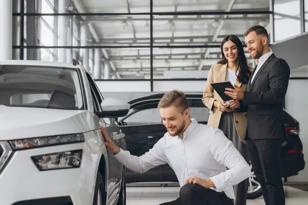 stock image Happy modern man inspecting new car in ar showroom and in background the manager of car showroom shows car catalog to woman