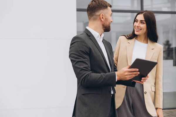 Продавец машин с бородой в сером костюме дает объяснения на планшете красивой молодой женщине в бежевом пиджаке возле автосалона