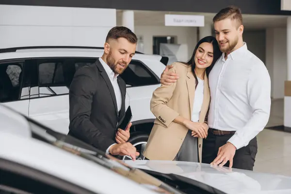 stock image A well-dressed salesman is presenting information to couple in a car dealership showroom. The couple appears interested as they stand between two new vehicles