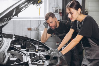 Otomobil servisi, tamirat, bakım konsepti. Tamirci benzin istasyonundaki arabayı kontrol ediyor. Çekici kadın ve sakallı mühendis araba aküsünü kontrol ediyor.
