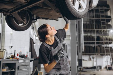 Araba tamirhanesinde araba tamircisi, gri üniformalı kadın tamirci tamirhanede araç bakımı yapıyor..