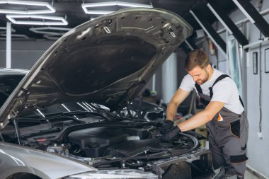 Yetenekli bir tamirci araba tamirhanesinde bir araba motorunu inceliyor, uzmanlık ve hassasiyet gösteriyor. Temel araçlar ve ekipmanlar arka planda görünür.