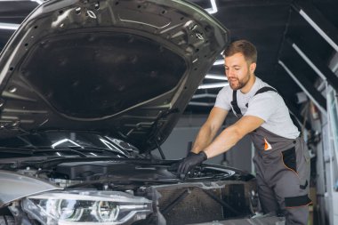 Tulum giyen mutlu oto tamircisi iyi aydınlatılmış bir garajda araba motorunda çalışıyor. Sahne, araç bakımında uzmanlık ve adanmışlığı yakalıyor.