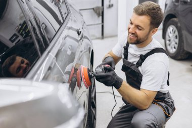 Garajda profesyonel bir araba parlatıcısı kullanarak araba bakım ve temizliği detaylarına titizlikle dikkat eden bir adamın yan görüntüsü..