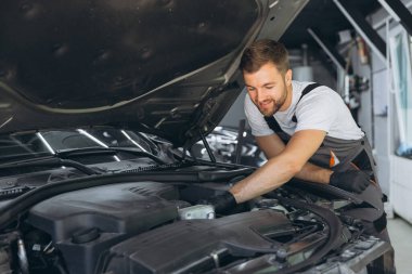 Bir oto tamir atölyesinde rutin araba bakımını yapan yetenekli bir tamirci, motor kontrolü ve servise odaklanıyor. Tamirci kaputun altında özenle çalışıyor..