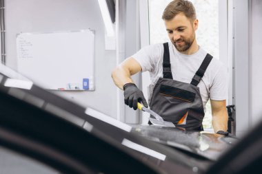 Adam araba kaputunu yıkıyor ve cam kaplama parlatma işlemini hazırlıyor. Erkek işçi garaja koruma filmi yerleştiriyor. Araç Ayrıntıları.