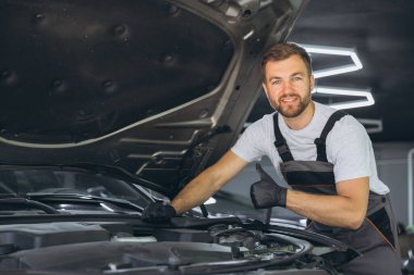 Üniformalı bir tamirci, bir oto tamir atölyesinde araba motorunu incelerken baş parmak hareketi yapıyor. Profesyonel hizmet ve araç bakımı.