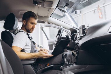 Otomotiv teknisyeni bilgisayarda tanılama yazılımı çalıştırıyor. Tamirci, oto servis garajında aracın tanılama testini yaparken arabada oturup dizüstü bilgisayarla çalışıyor..