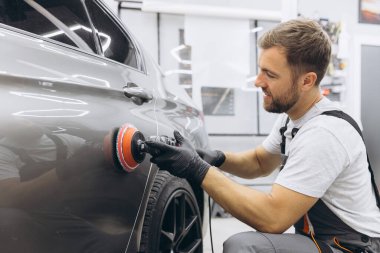 Garajda profesyonel bir araba parlatıcısı kullanarak araba bakım ve temizliği detaylarına titizlikle dikkat eden bir adamın yan görüntüsü..