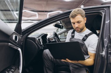 Otomotiv teknisyeni bilgisayarda tanılama yazılımı çalıştırıyor. Tamirci, oto servis garajında aracın tanılama testini yaparken arabada oturup dizüstü bilgisayarla çalışıyor..