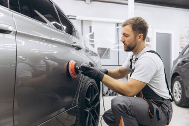 Garajda profesyonel bir araba parlatıcısı kullanarak araba bakım ve temizliği detaylarına titizlikle dikkat eden bir adamın yan görüntüsü..