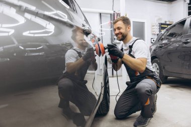 Garajda profesyonel bir araba parlatıcısı kullanarak araba bakım ve temizliği detaylarına titizlikle dikkat eden bir adamın yan görüntüsü..