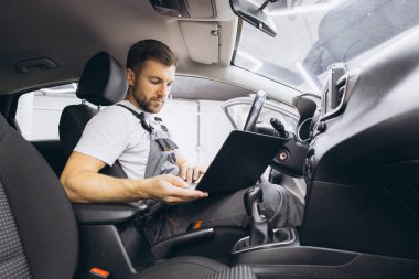 Otomotiv teknisyeni bilgisayarda tanılama yazılımı çalıştırıyor. Tamirci, oto servis garajında aracın tanılama testini yaparken arabada oturup dizüstü bilgisayarla çalışıyor..