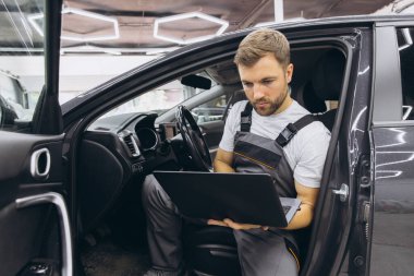 Otomotiv teknisyeni bilgisayarda tanılama yazılımı çalıştırıyor. Tamirci, oto servis garajında aracın tanılama testini yaparken arabada oturup dizüstü bilgisayarla çalışıyor..