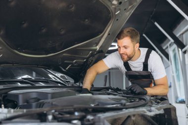 Üniformalı yetenekli bir tamirci modern bir oto tamirhanesinde araba motorunu incelerken gülümser. Uzmanlık ve adanmışlık gösterir..