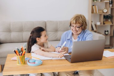 Mutlu büyükanne, kreş çocuğuna ev ödevi için yardım ediyor. Pozitif öğrenci kız ve büyükanne laptopta öğrenme kitabı okuyorlar, gülümsüyorlar, evde eğitimin tadını çıkarıyorlar.