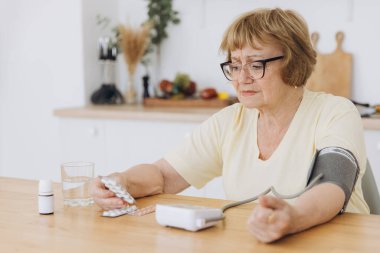 Yüksek tansiyonu olan yaşlı bir kadın evinde tansiyonunu ölçüyor. Üzgün yaşlı kadın kan basıncını tonometreyle ölçüyor. Günlük sağlık kontrolleri