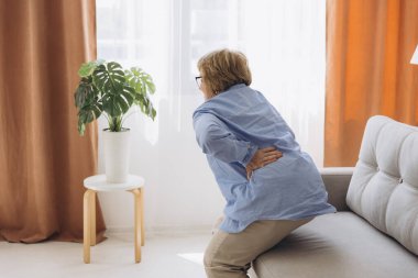 Unhappy old woman touching back, feeling pain, sitting on couch in living room, unhealthy middle aged senior female suffering from backache, spinal clipart